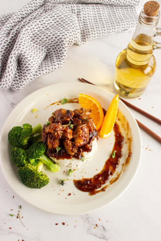 White plate with glazed meat, broccoli, and orange slice, with sauce; oil bottle and napkin in background.