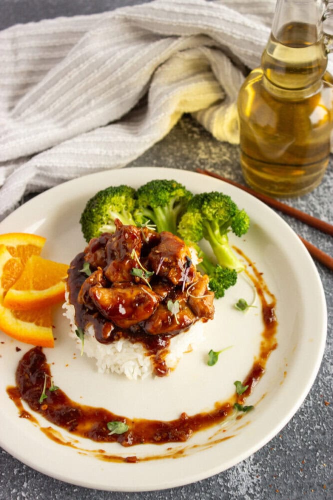 A plate of rice with glazed chicken, broccoli, orange slices, and sauce, with a bottle of oil in the background.
