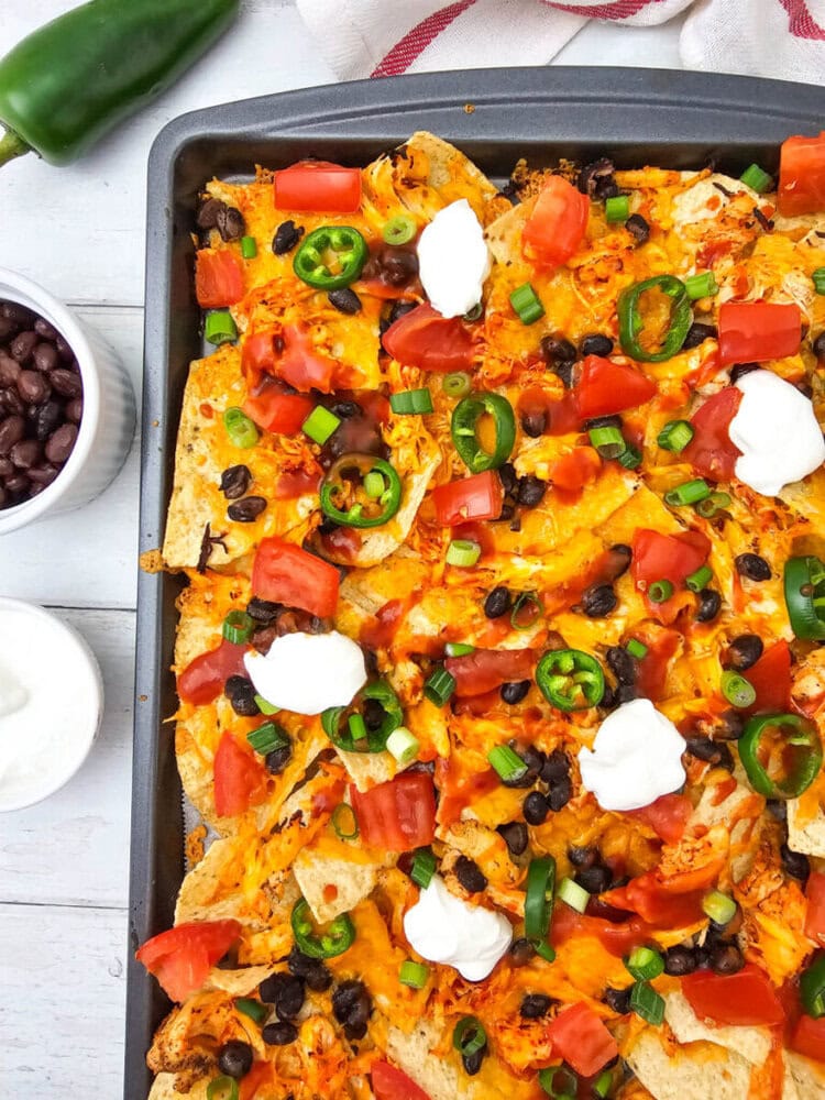 Sheet pan chicken nachos with cheese, black beans, peppers, jalape&ntilde;os, and sour cream on dark baking tray