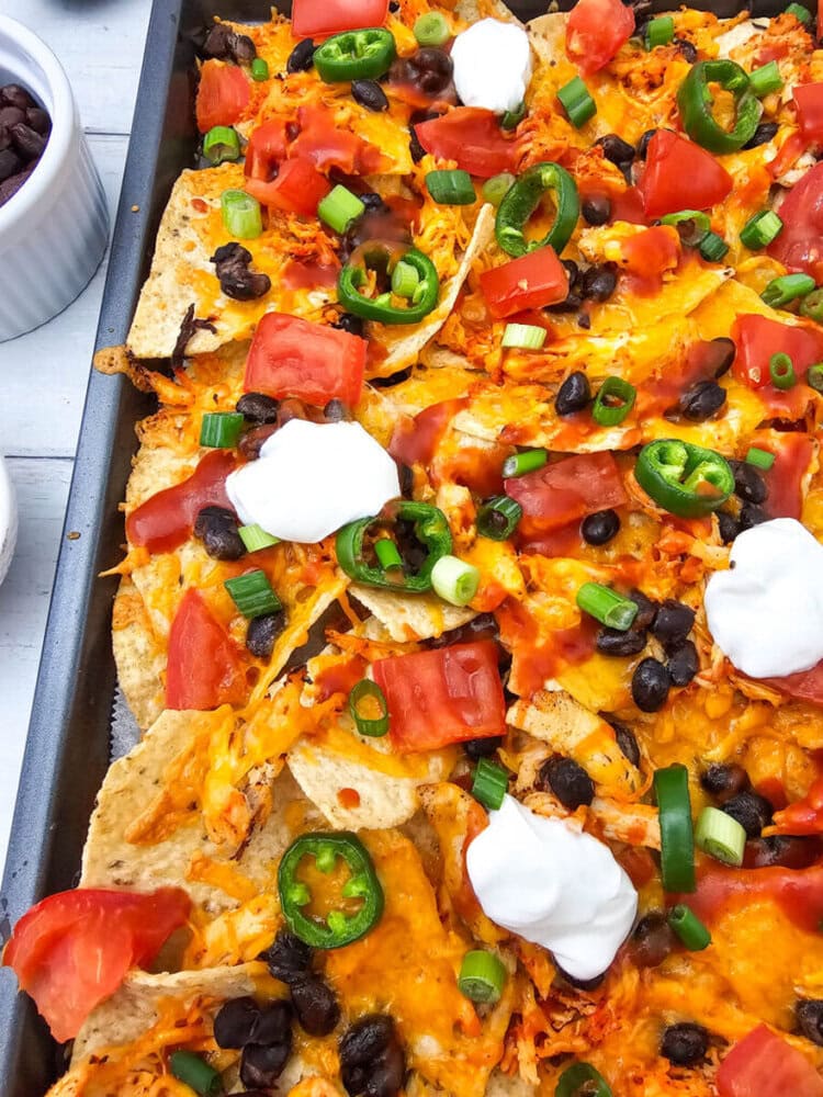 Sheet pan chicken nachos with cheese, black beans, jalape&ntilde;os, and sour cream on a baking tray
