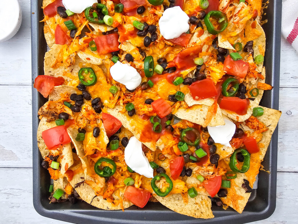 Sheet Pan Chicken Nachos with melted cheese, black beans, tomatoes, sour cream, and jalape&ntilde;os on a baking sheet