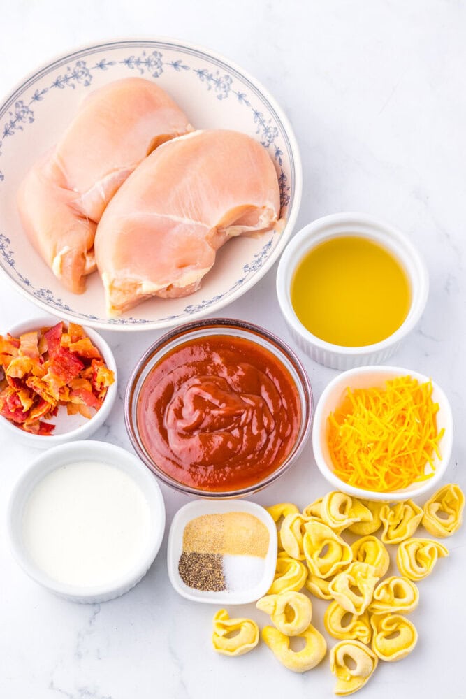 Ingredients for slow cooker BBQ chicken tortellini including raw chicken, BBQ sauce, cheese, tortellini, and seasonings arranged overhead