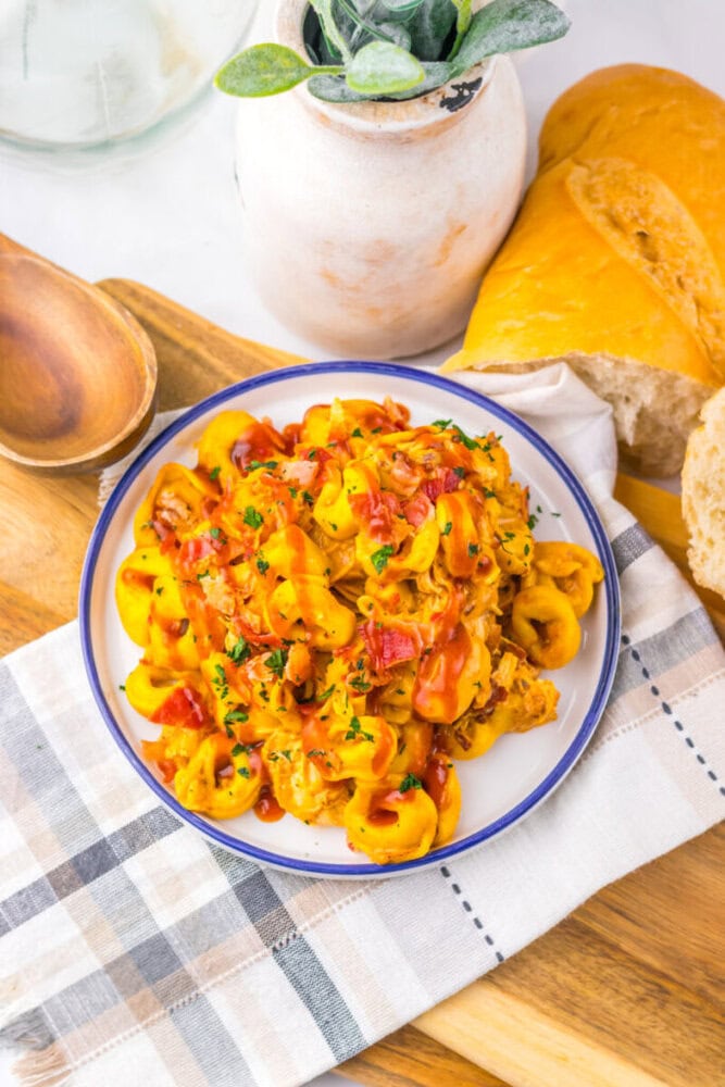 Slow cooker cheesy BBQ bacon chicken tortellini plated in white bowl with blue rim, garnished with fresh parsley and diced red peppers