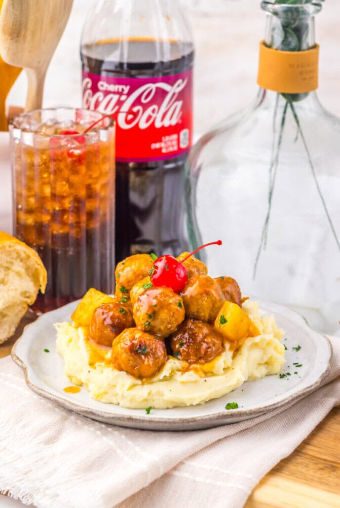 Slow cooker cherry cola pineapple BBQ meatballs served on mashed potatoes with cherry cola bottle and fresh herbs