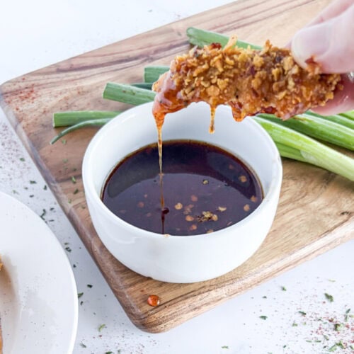 Crunchy baked chicken tender being dipped into chili infused honey sauce with green onions on wooden board