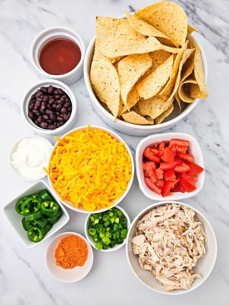 Sheet pan chicken nachos ingredients laid out including tortilla chips, shredded cheese, jalape&ntilde;os, salsa, sour cream, black beans, diced tomatoes, and shredded chicken