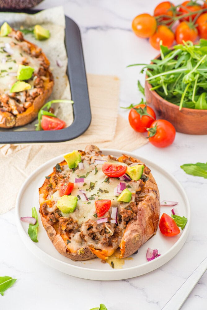 Stuffed sweet potato with ground meat, melted cheese, fresh toppings, and herbs on white plate