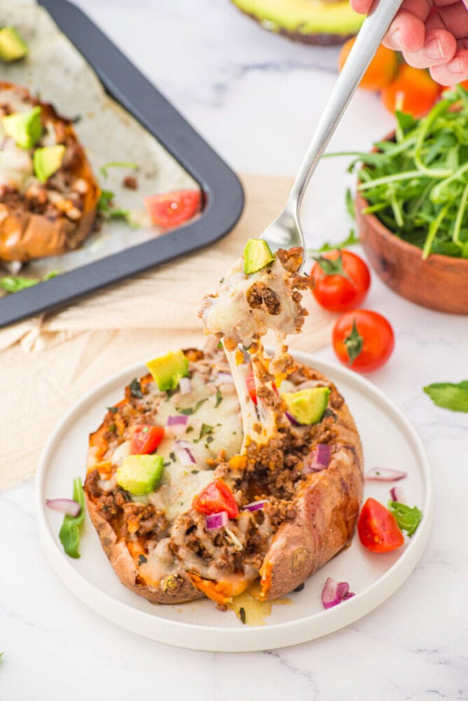 Stuffed sweet potato topped with melted cheese, diced peppers, onions, and arugula garnish on a white plate
