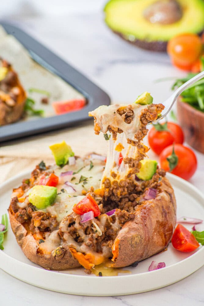Stuffed sweet potato with ground meat, melted cheese, and fresh vegetable toppings on a white plate