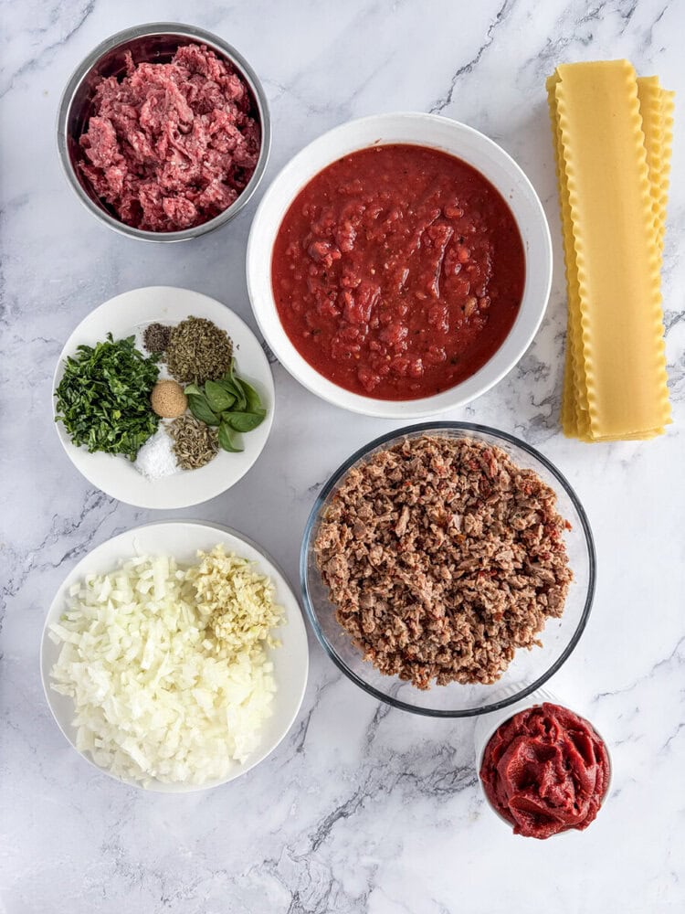 Lasagna ingredients prepped and laid out including ground beef, tomato sauce, pasta sheets, onions, and fresh herbs on marble countertop