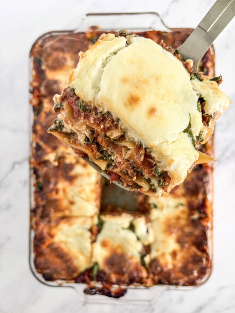 Baked 5 cheese lasagna with spinach and meat layers, slice being lifted with spatula showing melted cheese topping