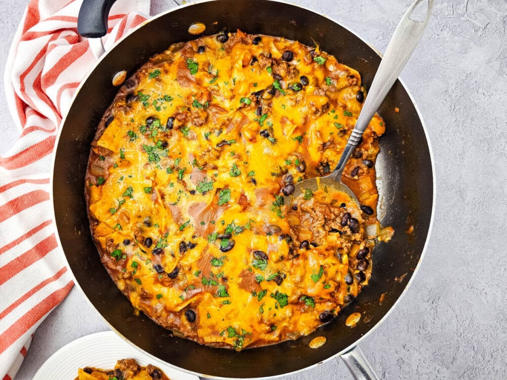 Beef enchiladas skillet with melted cheddar cheese, black beans, and cilantro garnish in a dark nonstick pan