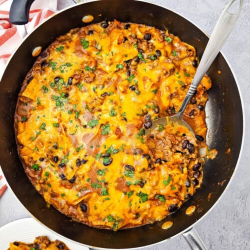 Beef enchiladas skillet with melted cheddar cheese, black beans, and cilantro garnish in a dark nonstick pan