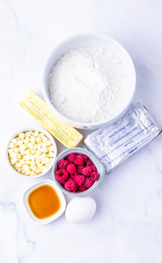 Ingredients for copycat Subway raspberry cheesecake cookies arranged overhead: flour, butter, white chocolate chips, raspberries, honey, and egg on marble background