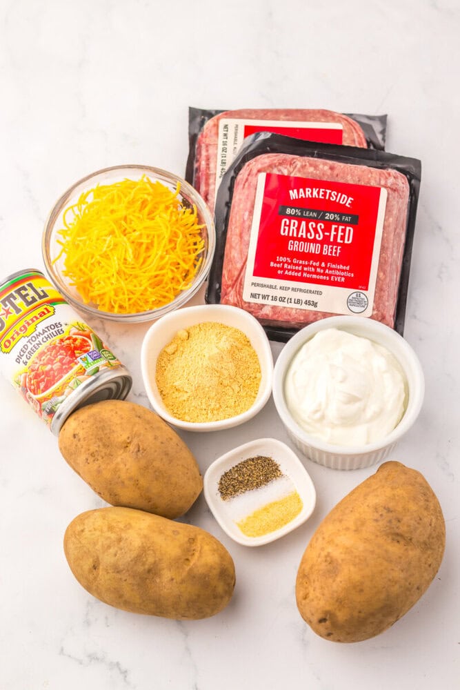 Ingredients for slow cooker cheesy taco potato casserole including ground beef, potatoes, shredded cheddar cheese, sour cream, diced tomatoes, and spices arranged on marble counter