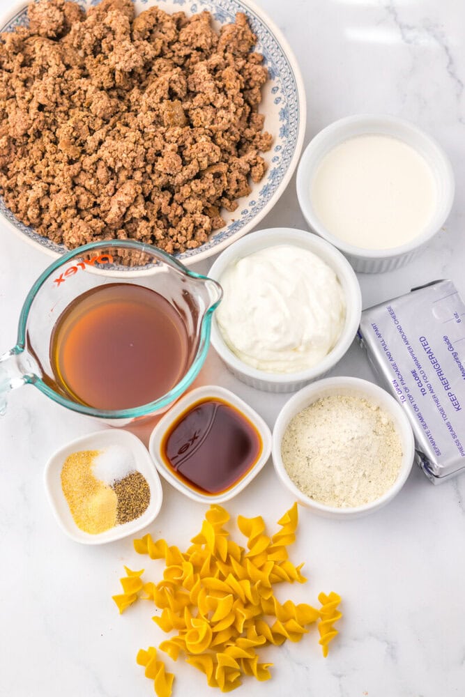 Ingredients for slow cooker creamy ranch beef and noodles including cooked ground beef, bow tie pasta, broth, sour cream, milk, and seasonings arranged on marble