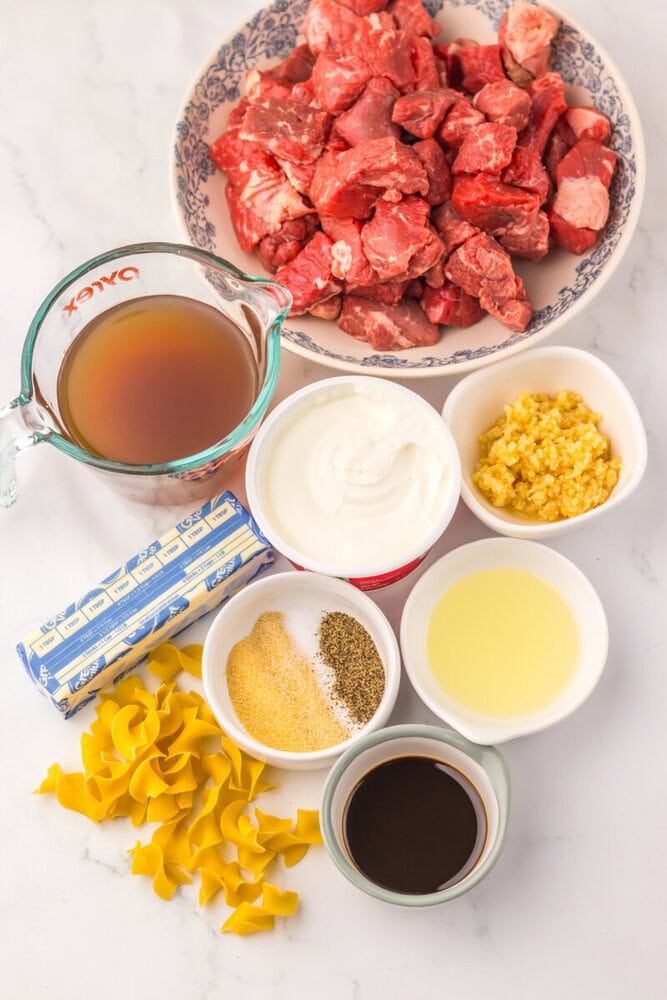 Slow cooker beef and noodles ingredients laid out: cubed beef, egg noodles, broth, sour cream, corn, spices, oil, and soy sauce