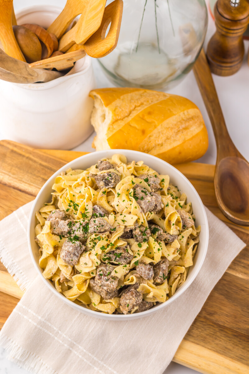 Slow cooker garlic butter beef and noodles in white bowl with parsley garnish, fresh bread and wooden utensils
