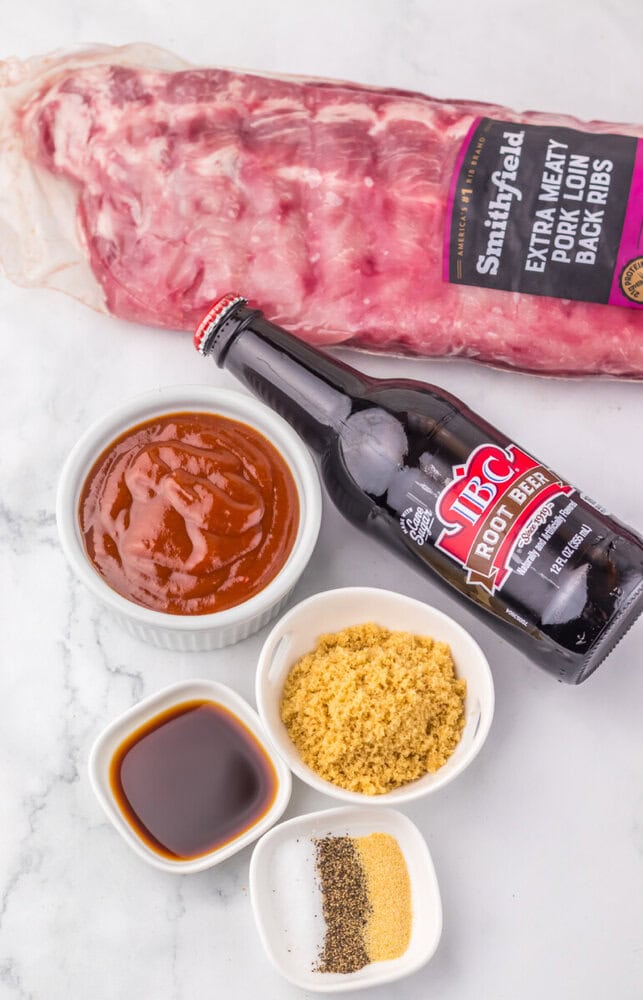 Slow cooker root beer BBQ ribs ingredients including raw pork ribs, BBQ sauce, root beer, brown sugar, and spices on marble surface
