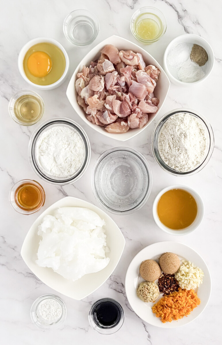 Orange chicken recipe ingredients arranged overhead including diced pork, flour, cornstarch, oils, and spices on marble counter