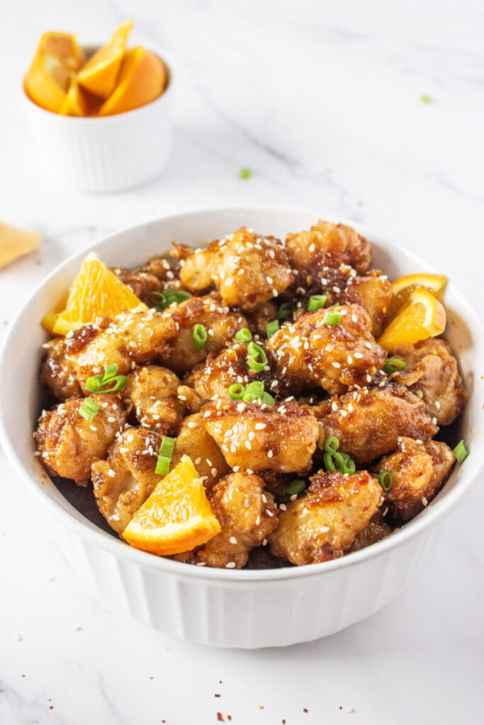 Glazed orange chicken with sesame seeds and scallions in white bowl with orange garnish
