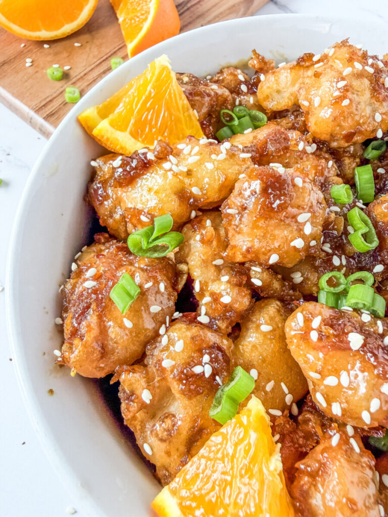 Orange chicken with sesame seeds and green onions served in a white bowl with fresh orange slices