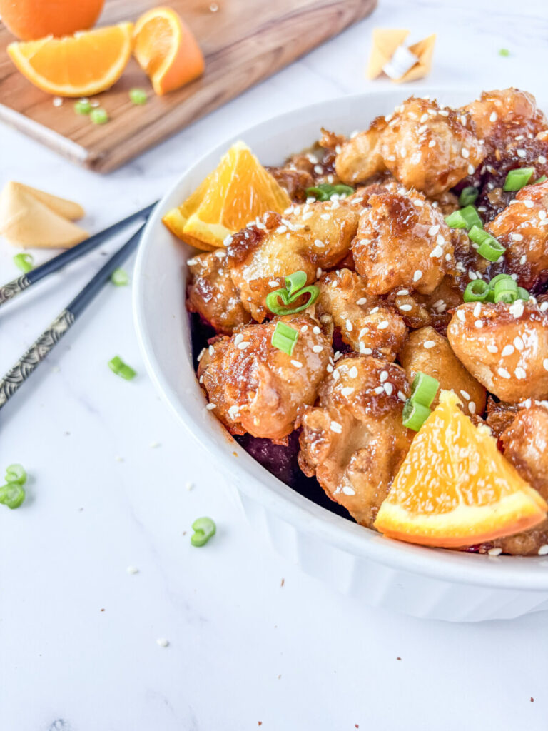 Orange chicken with sesame seeds and green onions garnish served in white bowl with orange slices