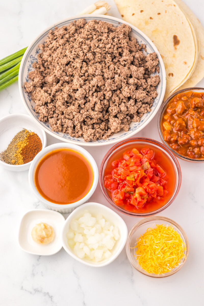 Slow cooker enchilada bake ingredients laid out in bowls including ground beef, tortillas, enchilada sauce, peppers, onions, and cheese