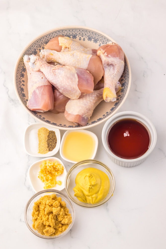 Slow cooker maple Dijon chicken legs ingredients: raw chicken pieces and bowls of spices, mustard, honey, and sauce on marble surface