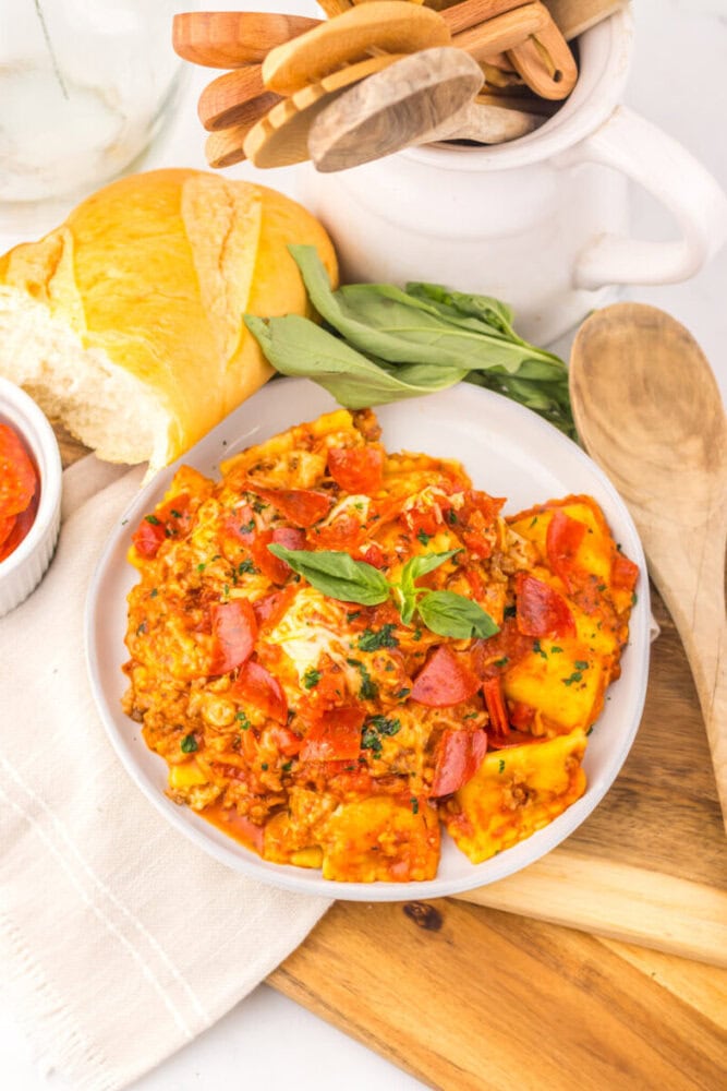 Slow cooker pizza ravioli served on white plate with pepperoni, basil, and tomatoes, overhead shot with fresh ingredients nearby