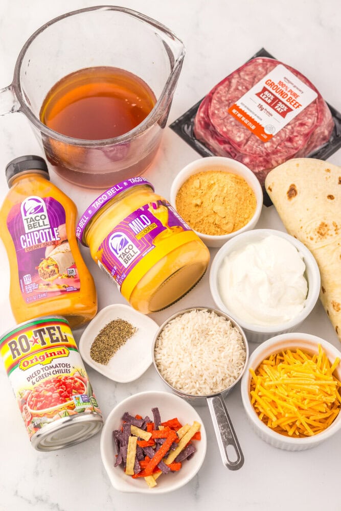 Slow cooker burrito casserole ingredients laid out including ground beef, tortillas, Taco Bell sauces, cheese, sour cream, and toppings