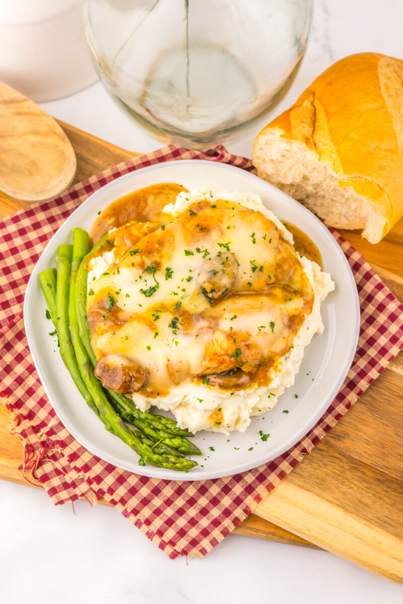 Slow cooker smothered chicken with melted cheese, mashed potatoes, and asparagus on white plate with bread and water glass