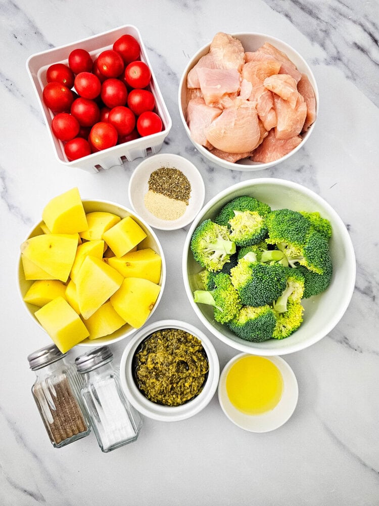 Sheet pan pesto chicken ingredients prepped and arranged overhead on marble counter including chicken, broccoli, potatoes, tomatoes, and seasonings