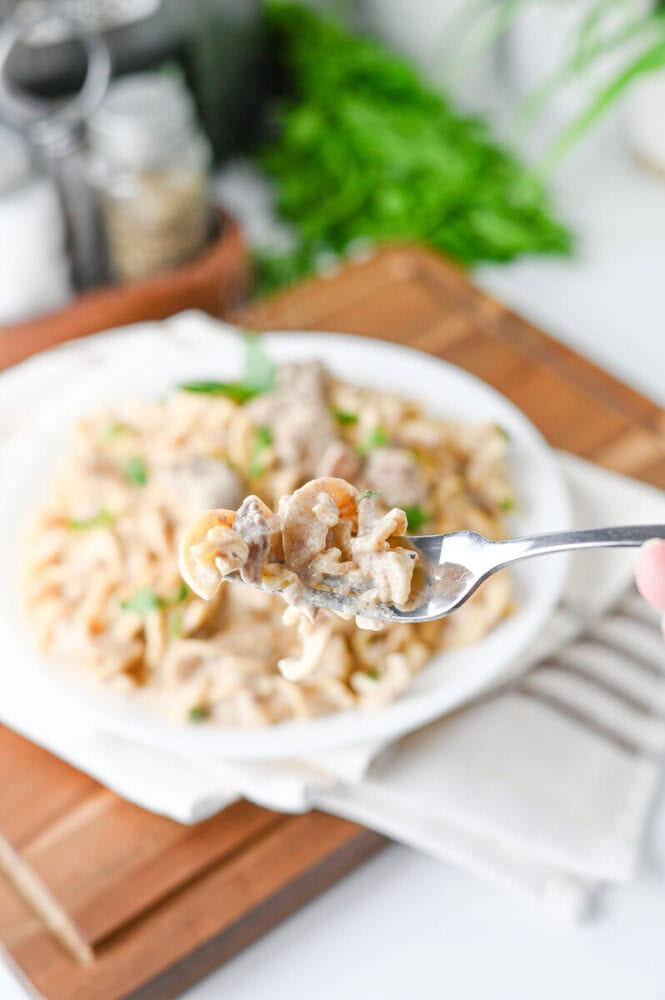 Slow cooker creamy beef and noodles on white plate with fork, garnished with fresh parsley