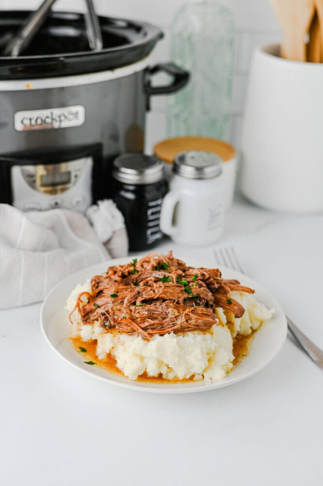 Slow Cooker Mississippi Pot Roast over mashed potatoes garnished with parsley, served on a white plate with slow cooker in background