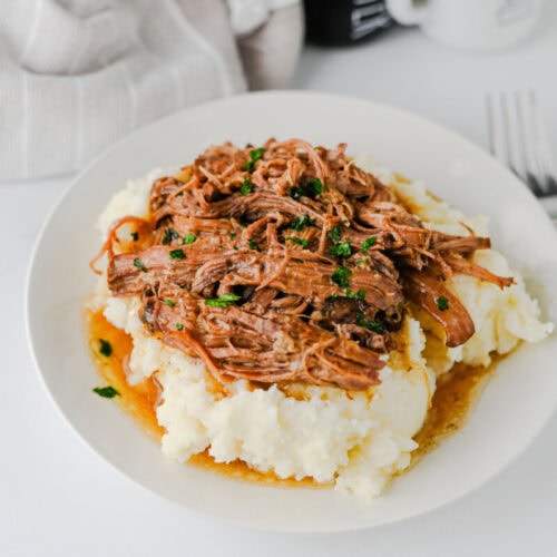 Slow Cooker Mississippi Pot Roast with shredded beef and peppers on mashed potatoes garnish with fresh herbs