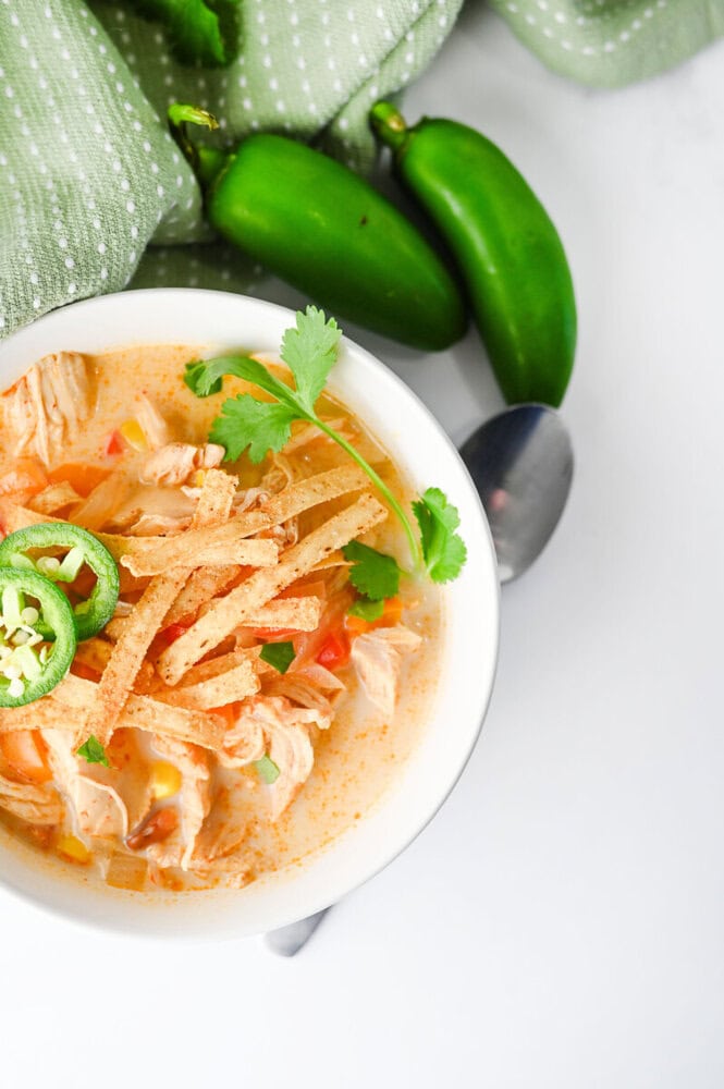 Slow cooker creamy chicken enchilada soup in white bowl with tortilla strips, jalapeños, and cilantro garnish