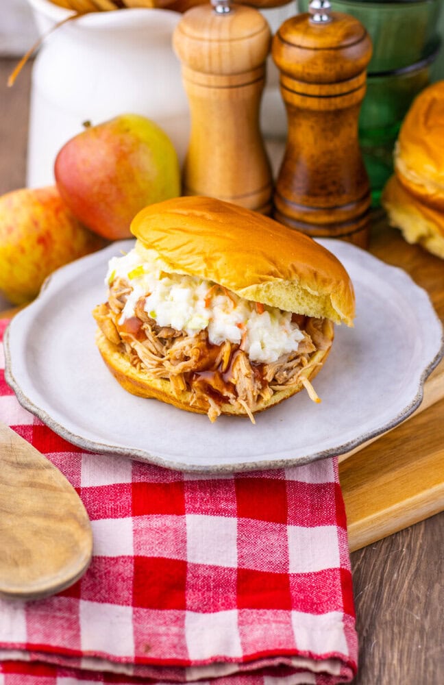 Apple cider BBQ pulled chicken sandwich with coleslaw on brioche bun, served on white plate with fresh apples and red gingham napkin
