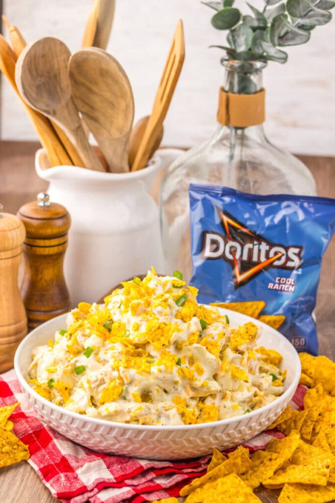 Cool Ranch Dorito Potato Salad in white bowl topped with crushed chips and garnish, surrounded by ingredients and styling props