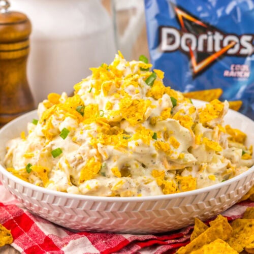 Cool Ranch Dorito potato salad in white bowl topped with crushed chips and garnish, surrounded by Doritos and table setting
