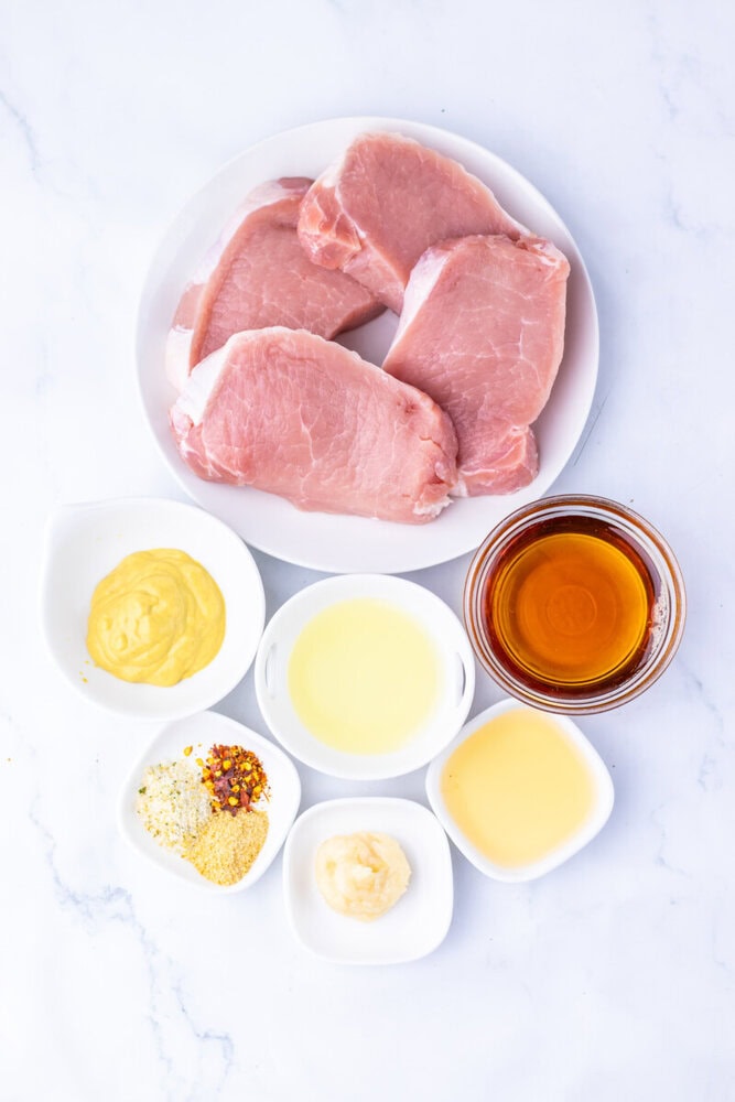 Maple Dijon pork chops ingredients laid out overhead including raw pork chops, Dijon mustard, maple syrup, honey, and spices on white bowls