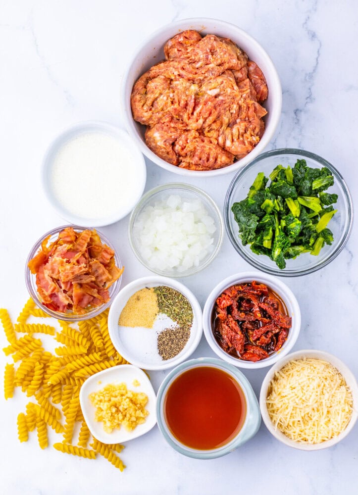 Zuppa Toscana pasta ingredients laid out in bowls including sausage, bacon, kale, pasta, broth, cheese, and seasonings on marble countertop