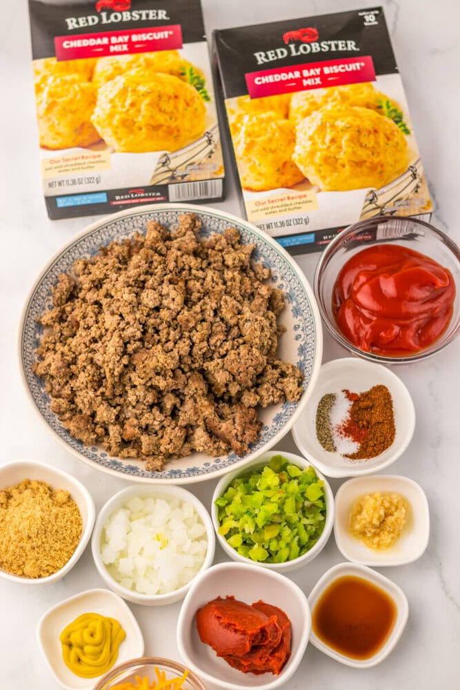 Slow cooker cheddar bay sloppy joe bake ingredients laid out overhead including ground beef, vegetables, spices, ketchup, and biscuit mix
