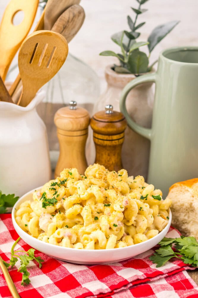 Creamy pasta with ham and cheese garnished with parsley in white bowl with wooden spoons and pepper mill in background