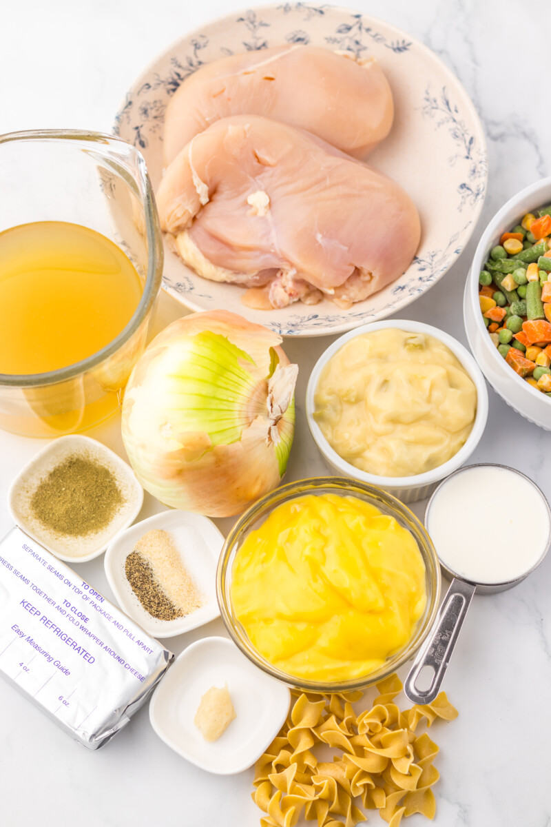 Slow cooker chicken pot pie pasta ingredients laid out including raw chicken, egg noodles, vegetables, broth, and seasonings