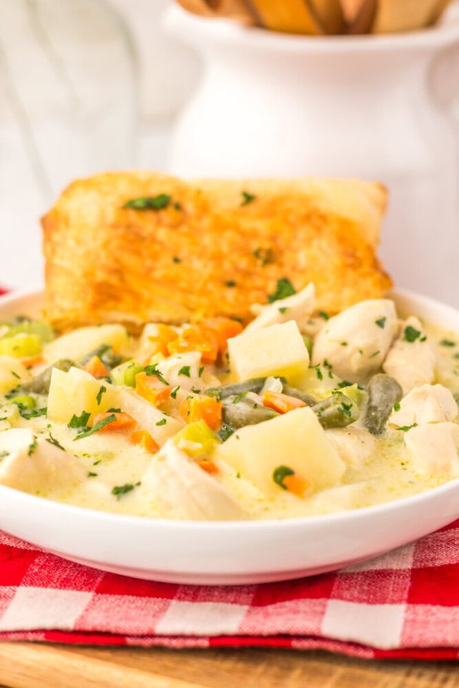 Slow cooker chicken pot pie soup in white bowl with vegetables, chicken, and biscuit on red checkered napkin