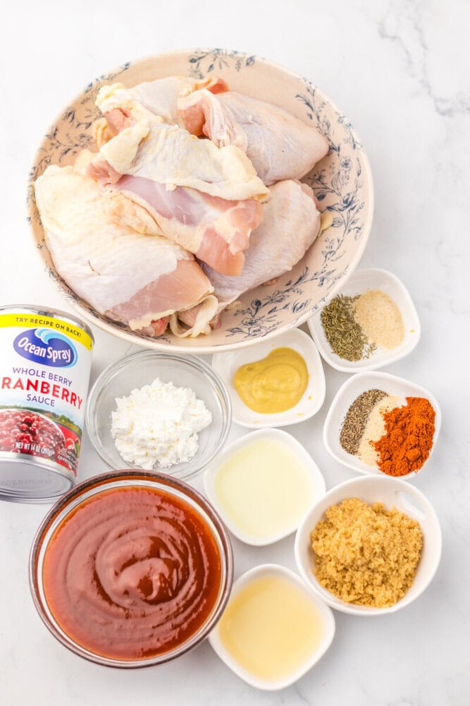 Slow cooker cranberry BBQ chicken ingredients including raw chicken, cranberry sauce, mustard, spices, and seasonings arranged overhead on marble surface