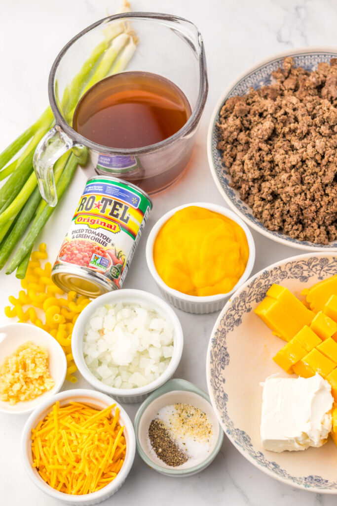 Slow Cooker Rotel Pasta ingredients laid out including ground beef, Rotel tomatoes, broth, pasta, cheese, diced peppers and onions in small bowls before cooking