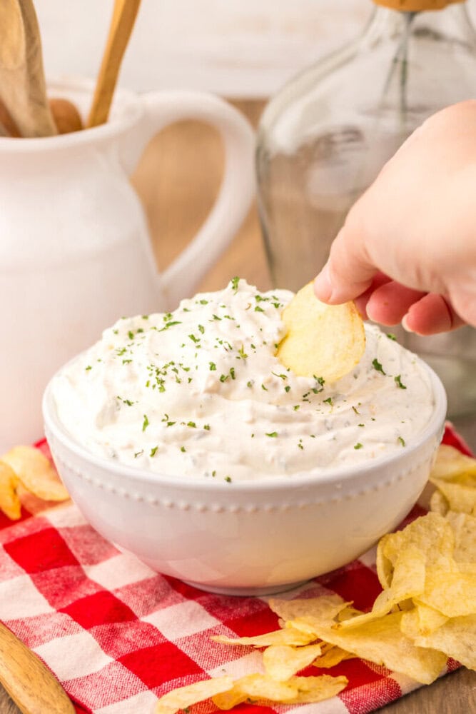 Creamy sour cream chip dip garnished with fresh herbs in a white bowl with potato chips and a hand dipping a chip