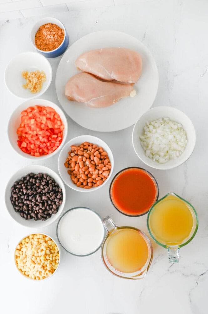 Slow cooker creamy chicken enchilada soup ingredients laid out in bowls including chicken, beans, corn, peppers, onions, and enchilada sauce